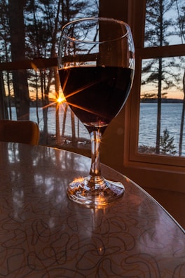 A wine glass filled with red wine is positioned on a table indoors, with the backdrop of a scenic sunset over a body of water. Sunlight streams through the trees and the window, casting reflections and creating a warm ambiance. The surrounding area is tranquil, with the silhouette of trees against the colorful sky.
