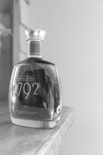 A bottle of 1792 small batch Kentucky straight bourbon whiskey sits on a wooden surface. The bottle has a distinct curvy shape with a glass stopper and is partially full. The image is in black and white, creating a classic and elegant feel.