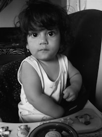 A young child with curly hair is gazing intently at the camera. The child's face is expressive, and they are wearing a sleeveless shirt. The setting includes a baby walker tray with small toys, such as a steering wheel and animal figures.