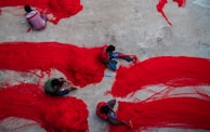 Several individuals are working on spreading and organizing large red nets on a concrete surface. The bright red of the nets contrasts with the dull grey of the ground. The people are seated closely and appear focused on their task.