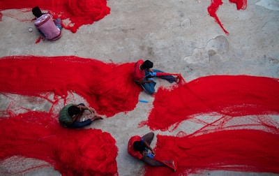 Several individuals are working on spreading and organizing large red nets on a concrete surface. The bright red of the nets contrasts with the dull grey of the ground. The people are seated closely and appear focused on their task.