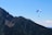 A paraglider with a blue and white canopy soars high above a mountain range with lush green forests. The sky is clear and blue, and a few clouds are visible in the distance.