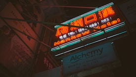 A vibrant neon sign with bright orange and teal colors, featuring both Chinese and English text advertising a hotel. Below it is another sign for Alchemy, possibly a business or service. The scene is set at night, with a dark background and glimpses of nearby buildings adorned with posters.