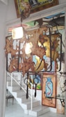 An artistic chandelier made from rusted metal elements hangs from the ceiling. It casts an ambient light in a room with brightly colored wall art that includes abstract and graffiti-like designs. A staircase with white steps and a decorative railing are visible in the background, along with a small table.