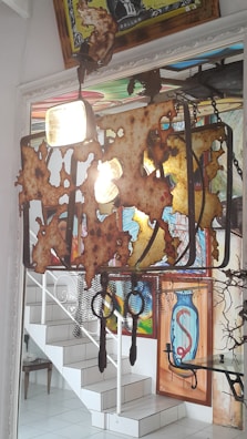 An artistic chandelier made from rusted metal elements hangs from the ceiling. It casts an ambient light in a room with brightly colored wall art that includes abstract and graffiti-like designs. A staircase with white steps and a decorative railing are visible in the background, along with a small table.