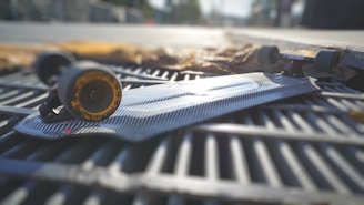 A skateboard with a carbon-fiber patterned deck is lying on a metal grate. The wheels are visible with a yellow outer rim, and the sunlight casts reflections on the surface. The background is slightly blurred, creating a shallow depth of field effect.
