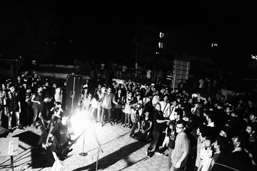 A lively outdoor concert with a large crowd gathered around a stage. The scene is captured in black and white, adding a dramatic and classic feel. The band is performing with visible musical instruments and a microphone. Attendees are diverse in appearance and appear to be enjoying the performance.