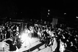 A lively outdoor concert with a large crowd gathered around a stage. The scene is captured in black and white, adding a dramatic and classic feel. The band is performing with visible musical instruments and a microphone. Attendees are diverse in appearance and appear to be enjoying the performance.