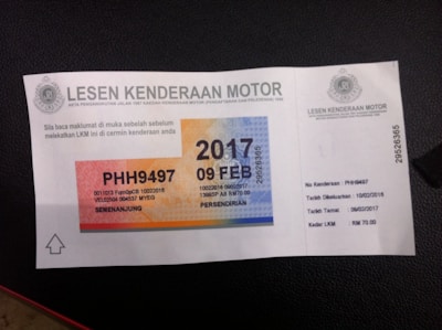 The image features a Malaysian Motor Vehicle License document with a colorful design, displaying the text 'LESEN KENDERAAN MOTOR' along with a vehicle registration number and expiration date. The license has a gradient of orange, blue, and yellow and includes intricate security patterns.