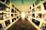 A long, narrow corridor with multiple rows of mausoleum niches lining both sides. The walls are covered with plaques and floral tributes, creating a solemn atmosphere. The perspective draws the eye towards the light at the end of the corridor.