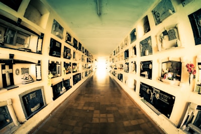 A long, narrow corridor with multiple rows of mausoleum niches lining both sides. The walls are covered with plaques and floral tributes, creating a solemn atmosphere. The perspective draws the eye towards the light at the end of the corridor.