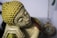 A close-up of a decorative Buddha sculpture with a textured yellow head and serene facial expression. In the background, a hanging pocket watch is visible, slightly blurred.
