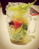 A clear mason jar filled with various fresh vegetables, including lettuce, is placed on a light-colored surface. The jar contains layers of green, red, and white produce, adding a vibrant mix of colors. A smartphone with a red case and a dark glass are visible in the background.