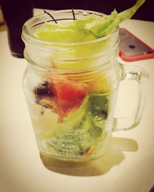 A clear mason jar filled with various fresh vegetables, including lettuce, is placed on a light-colored surface. The jar contains layers of green, red, and white produce, adding a vibrant mix of colors. A smartphone with a red case and a dark glass are visible in the background.