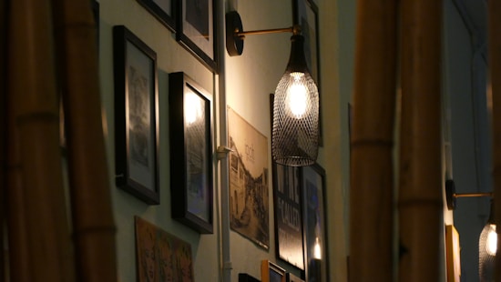 A dimly lit interior wall featuring a row of framed photographs and artwork, alongside modern, softly glowing wall lamps with mesh-like coverings. Vertical wooden beams partially frame the view.