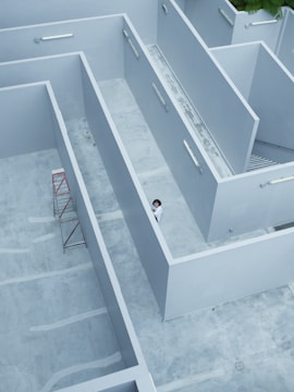 Aerial view of a minimalist architectural structure with clean, geometric lines and a color palette dominated by light grey. A person in white clothing is standing at the intersection of the walls, adding a sense of scale and human presence. A red scaffolding is positioned against one of the interior walls. The area appears to be a rooftop or courtyard space with a concrete floor.