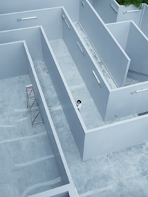 Aerial view of a minimalist architectural structure with clean, geometric lines and a color palette dominated by light grey. A person in white clothing is standing at the intersection of the walls, adding a sense of scale and human presence. A red scaffolding is positioned against one of the interior walls. The area appears to be a rooftop or courtyard space with a concrete floor.
