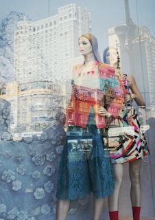 Mannequins in a store display window with reflections of an urban cityscape superimposed. The mannequins are dressed in colorful, fashionable clothing against a backdrop featuring a rose pattern.