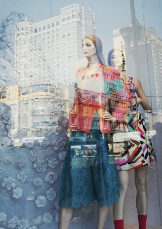 Mannequins in a store display window with reflections of an urban cityscape superimposed. The mannequins are dressed in colorful, fashionable clothing against a backdrop featuring a rose pattern.