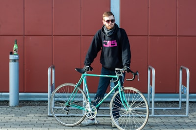 A person wearing sunglasses, a black hoodie with pink lettering, and a scarf stands next to a teal road bicycle. The individual is positioned in front of a red wall with rectangular panels and is standing on a brick pavement. Nearby, a tall, slender bollard supports an upright bottle.