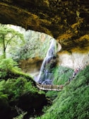 A scenic view inside a lush green cave-like structure with a waterfall cascading down a rocky wall. A wooden walkway with railings spans the scene, featuring a group of people observing the natural beauty around them. Stalactites hang from the ceiling and dense foliage surrounds the area, enhancing the serene and natural atmosphere.