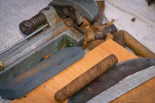 An assortment of cigars and a cigar cutter are placed on a wooden surface. The cutter displays details of machinery with metallic components and a spring mechanism. The cigars have a rich brown color, indicating different stages of the rolling process.