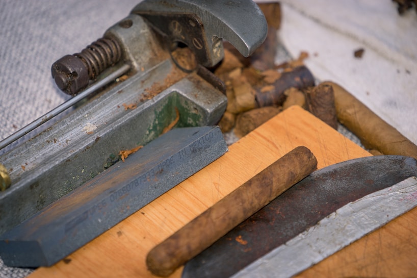 An assortment of cigars and a cigar cutter are placed on a wooden surface. The cutter displays details of machinery with metallic components and a spring mechanism. The cigars have a rich brown color, indicating different stages of the rolling process.