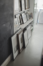 Several framed pictures and photographs are stacked and propped up against a wall and on shelves. The background includes a chalkboard surface on the left side and a glass door with a metal handle to the right. The frames are of various sizes and colors, some leaning against one another.