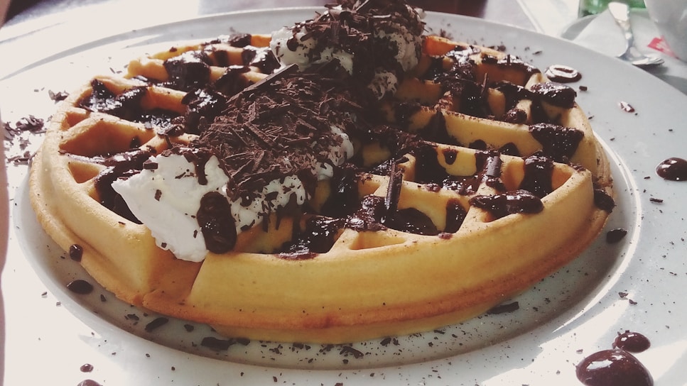 A delicious waffle topped with whipped cream, chocolate sauce, and chocolate shavings, served on a white plate with scattered chocolate droplets around it.