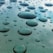 Numerous droplets of water are scattered across a smooth surface, reflecting ambient light. The droplets vary in size, creating a textured appearance, while the background is a glossy mix of blue and green hues.