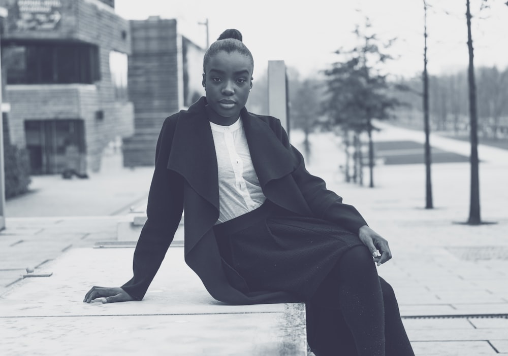 grayscale photo of woman sitting on concrete pavement near building