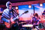 A live music performance with a guitarist on the left wearing a patterned poncho and a beanie, playing an electric guitar. To the right, a drummer is energetically playing a drum set. The backdrop features a desert landscape with cacti, creating a vibrant and dynamic stage environment.