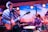 A live music performance with a guitarist on the left wearing a patterned poncho and a beanie, playing an electric guitar. To the right, a drummer is energetically playing a drum set. The backdrop features a desert landscape with cacti, creating a vibrant and dynamic stage environment.