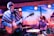A live music performance with a guitarist on the left wearing a patterned poncho and a beanie, playing an electric guitar. To the right, a drummer is energetically playing a drum set. The backdrop features a desert landscape with cacti, creating a vibrant and dynamic stage environment.