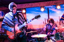 A live music performance with a guitarist on the left wearing a patterned poncho and a beanie, playing an electric guitar. To the right, a drummer is energetically playing a drum set. The backdrop features a desert landscape with cacti, creating a vibrant and dynamic stage environment.