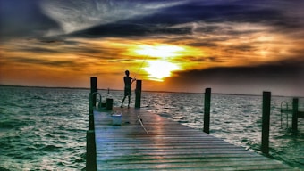 A solitary figure stands on a wooden pier extending into a large body of water, fishing rod in hand against a dramatic sunset. The sky is filled with vibrant hues of orange, yellow, and dark clouds, casting reflections on the choppy waves surrounding the pier.