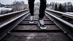 person wearing pair of black-and-white Vans Old Skool shoes walking on brown train tracks during winter