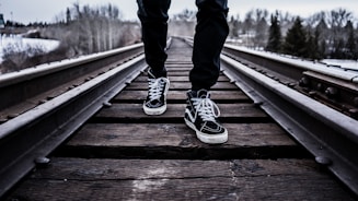 person wearing pair of black-and-white Vans Old Skool shoes walking on brown train tracks during winter