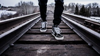 A person wearing black pants and black high-top sneakers with white laces is walking on a wooden railway track. The scene is set in a wintry landscape with sparse trees and snow visible in the background.