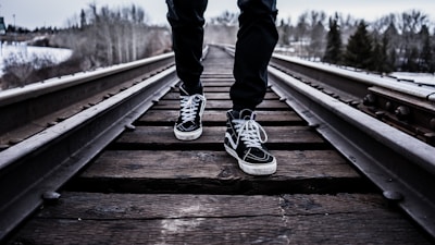 A person wearing black pants and black high-top sneakers with white laces is walking on a wooden railway track. The scene is set in a wintry landscape with sparse trees and snow visible in the background.