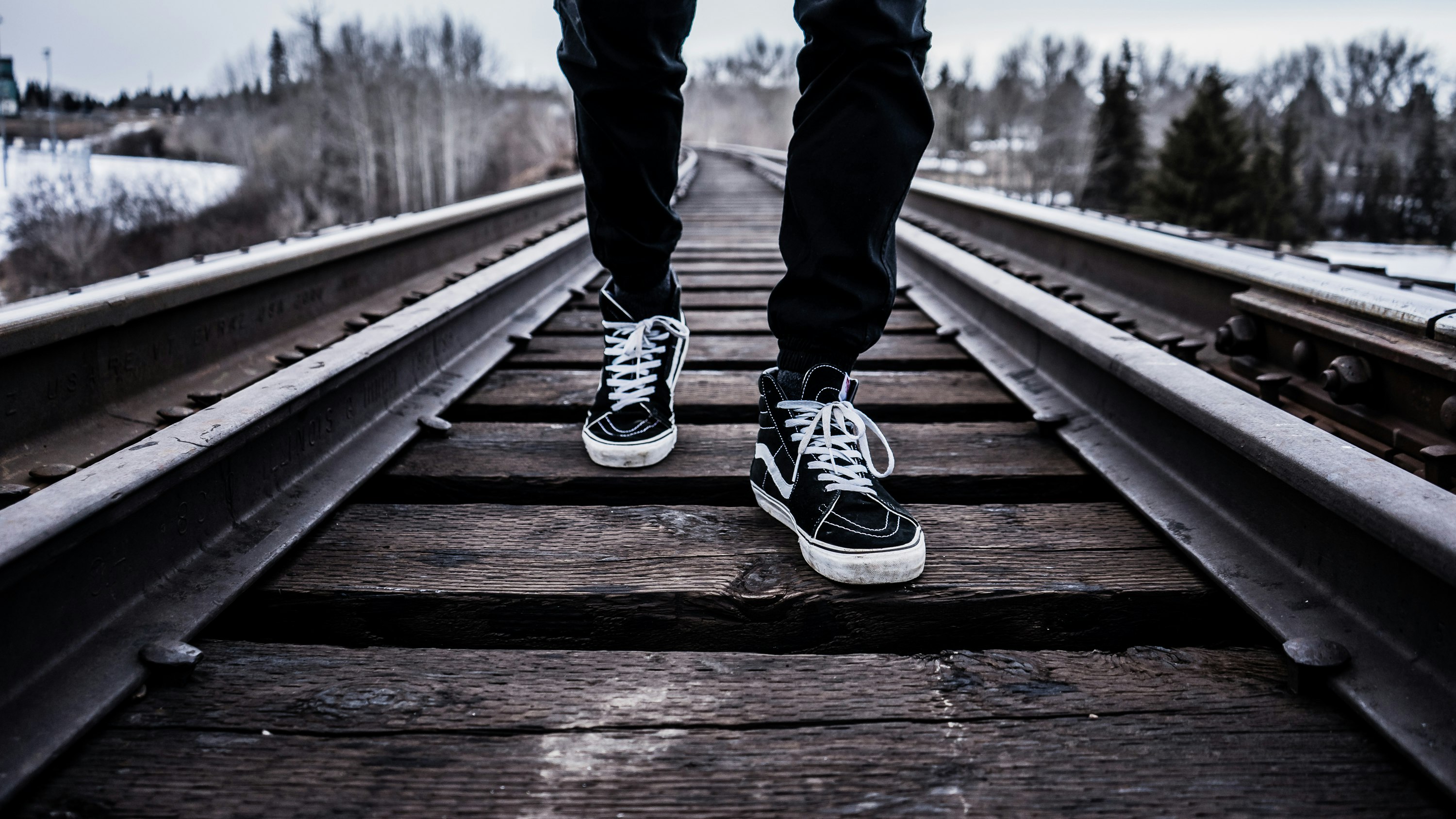 person wearing pair of black-and-white Vans Old Skool shoes walking on brown train tracks during winter