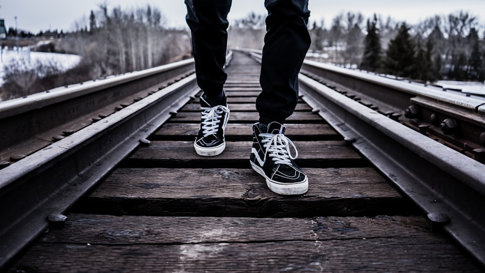 persona con un par de zapatos Vans Old Skool blancos y negros caminando sobre las vías del tren marrón durante el invierno