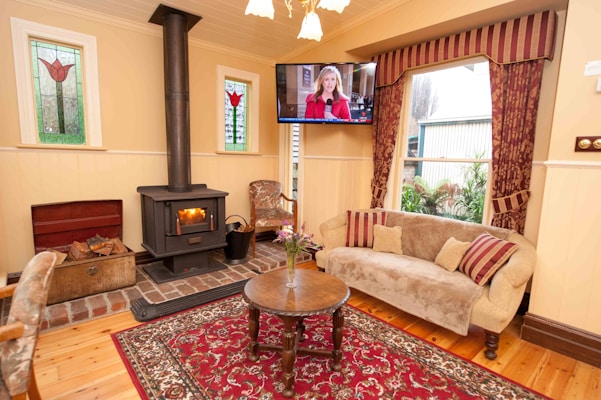 A cozy living room with a wooden floor and a richly patterned red rug. In the corner, a wood-burning stove is lit, illuminating the space with a warm glow. Next to it is a basket filled with logs. A plush sofa with decorative striped pillows adds comfort, and a round wooden table with a vase of flowers enhances the homely atmosphere. Stained glass windows featuring floral designs bring in natural light, while a TV is mounted above. The room is decorated with elegant drapes and classic furniture.