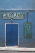 A blue facade with a large blue door and two green signs featuring white, bold text reading 'ANTIQUITES'. The signs seem to advertise buying, selling, and exchanging antiques, with additional contact information visible. A single wall-mounted light fixture is centered above the door.