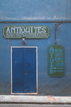 A blue facade with a large blue door and two green signs featuring white, bold text reading 'ANTIQUITES'. The signs seem to advertise buying, selling, and exchanging antiques, with additional contact information visible. A single wall-mounted light fixture is centered above the door.