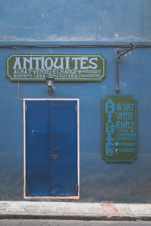 A blue facade with a large blue door and two green signs featuring white, bold text reading 'ANTIQUITES'. The signs seem to advertise buying, selling, and exchanging antiques, with additional contact information visible. A single wall-mounted light fixture is centered above the door.