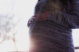 A person is gently holding their pregnant belly, wearing a grey knit sweater. Soft, natural light filters in from the background, creating a serene and intimate atmosphere.