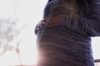 A person is gently holding their pregnant belly, wearing a grey knit sweater. Soft, natural light filters in from the background, creating a serene and intimate atmosphere.