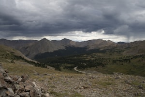 A mountainous landscape with rugged terrain and a winding road leading into a dense forest. The sky is overcast with dark, dramatic clouds that hint at an impending storm. Sparse vegetation covers the rocky ground in the foreground.