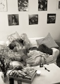 A cozy living room setting with a white sofa covered in a textured blanket and a black-and-white patterned pillow. On the coffee table, there are magazines, a candle, and a small decorative object. A vase with flowers is positioned at the edge of the table. The wall is adorned with several black-and-white posters and photographs, contributing to a vintage ambiance. The double exposure effect gives the image a dreamy, surreal vibe.